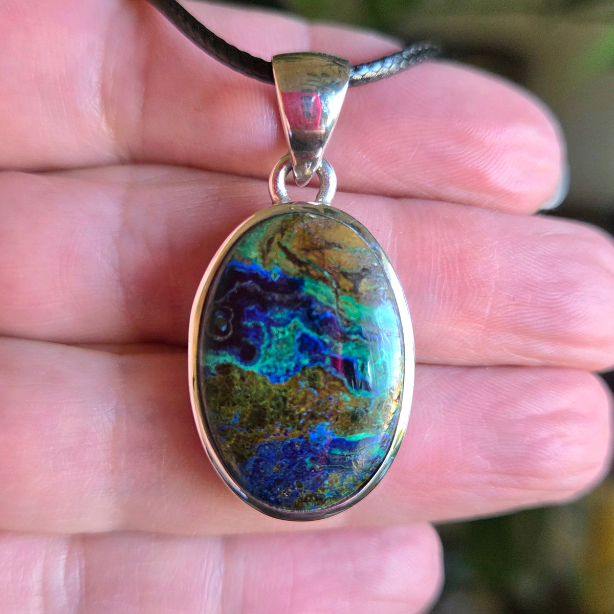 An oval-shaped Azurite Malachite pendant held in a person's hand, with a multicolored stone predominantly in shades of blue and green, set in a silver-colored frame.