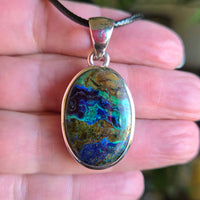 An oval-shaped Azurite Malachite pendant held in a person's hand, with a multicolored stone predominantly in shades of blue and green, set in a silver-colored frame.