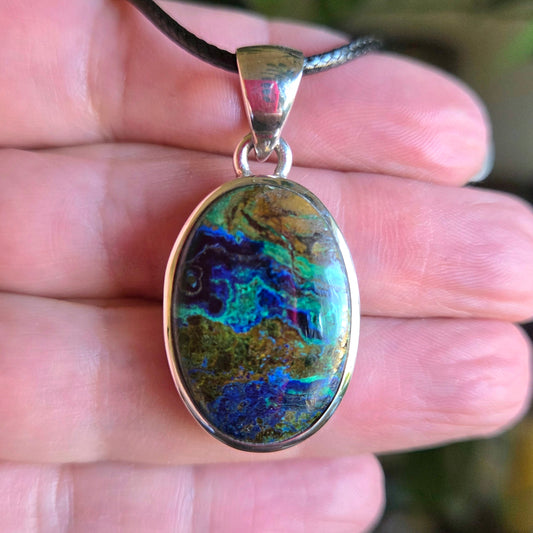 An oval-shaped Azurite Malachite pendant held in a person's hand, with a multicolored stone predominantly in shades of blue and green, set in a silver-colored frame.