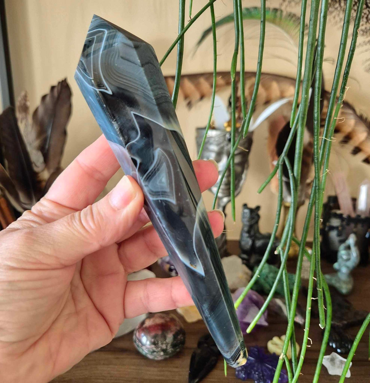A hand holding a Black Agate sceptre with smoky swirl patterns, displayed in front of a assortment of feathered ornaments and crystals.