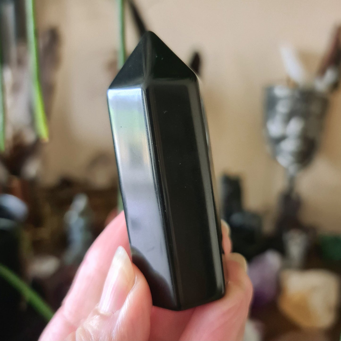 A small, black obsidian crystal generator held in a person's hand, with a blurred background featuring other crystals and spiritual artifacts.
