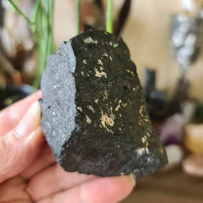 A large black tourmaline crystal stone held in a person's hand, with a blurred background suggesting a spiritual or metaphysical context.