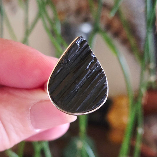 A hand holding a black tourmaline ring with a silver band, showing the textured surface of the stone.