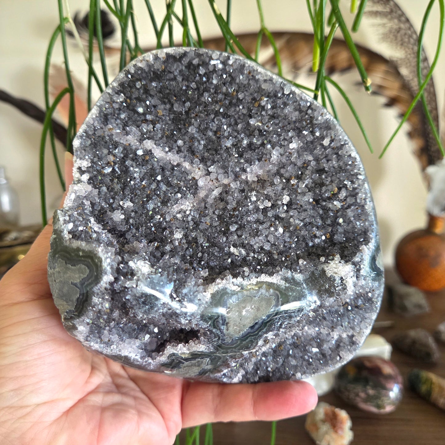 Hand holding a large crystal rock with a textured surface, surrounded by plants and other stones.
