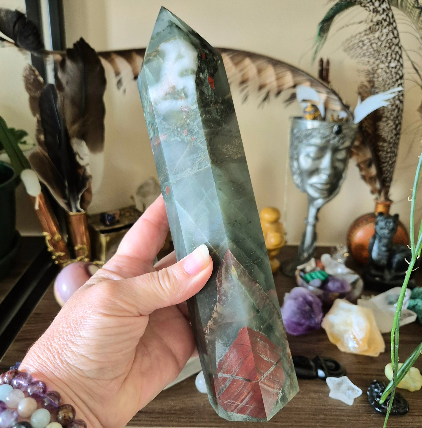 A hand holding a polished bloodstone crystal with a reddish and greenish color, possibly a tower-shaped variant.