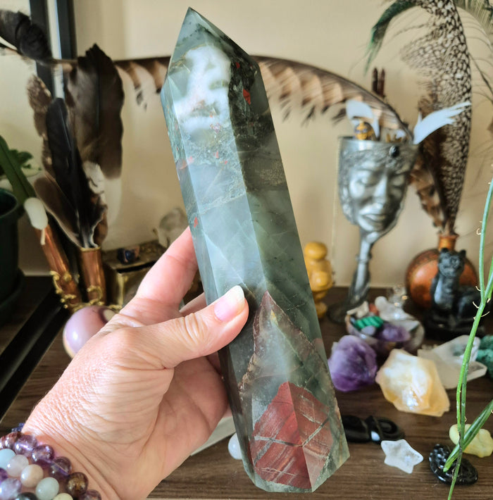 A hand holding a polished bloodstone crystal with a reddish and greenish color, possibly a tower-shaped variant.