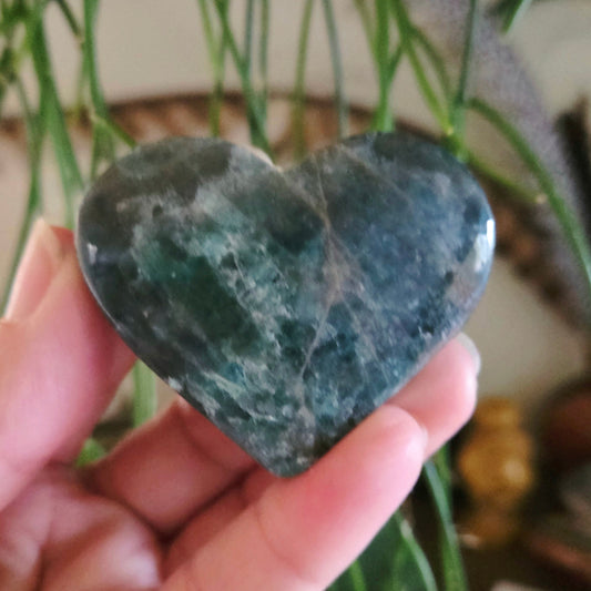 A heart-shaped Blue Apatite stone held in a person's hand, with a greenish-blue color and natural patterns.