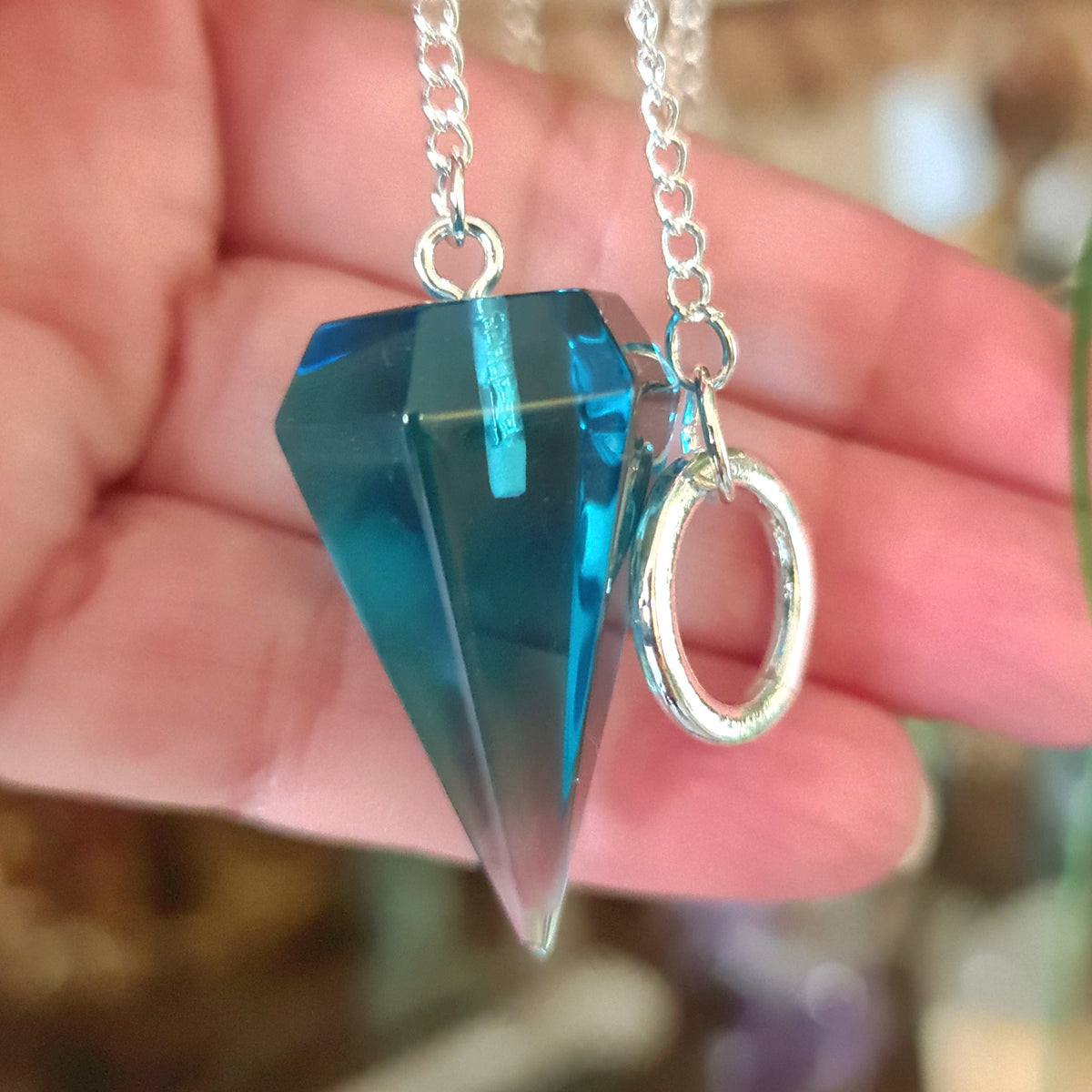A blue obsidian pendulum with a silver-colored ring at the bottom, held in a person's hand.