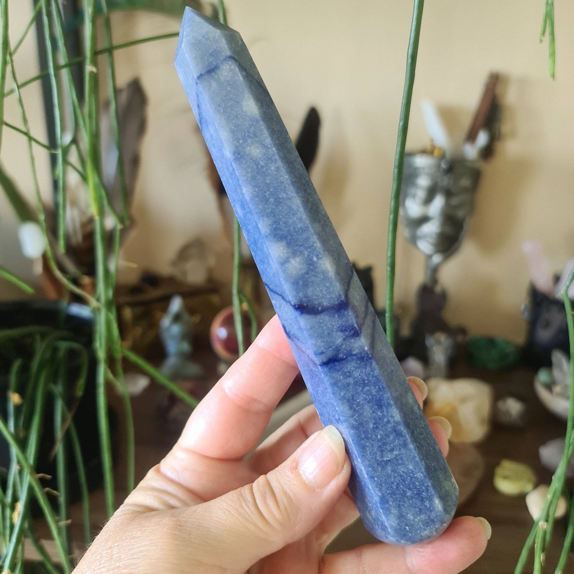 A hand holding a blue quartz crystal wand with a plant and various crystal figures in the background.