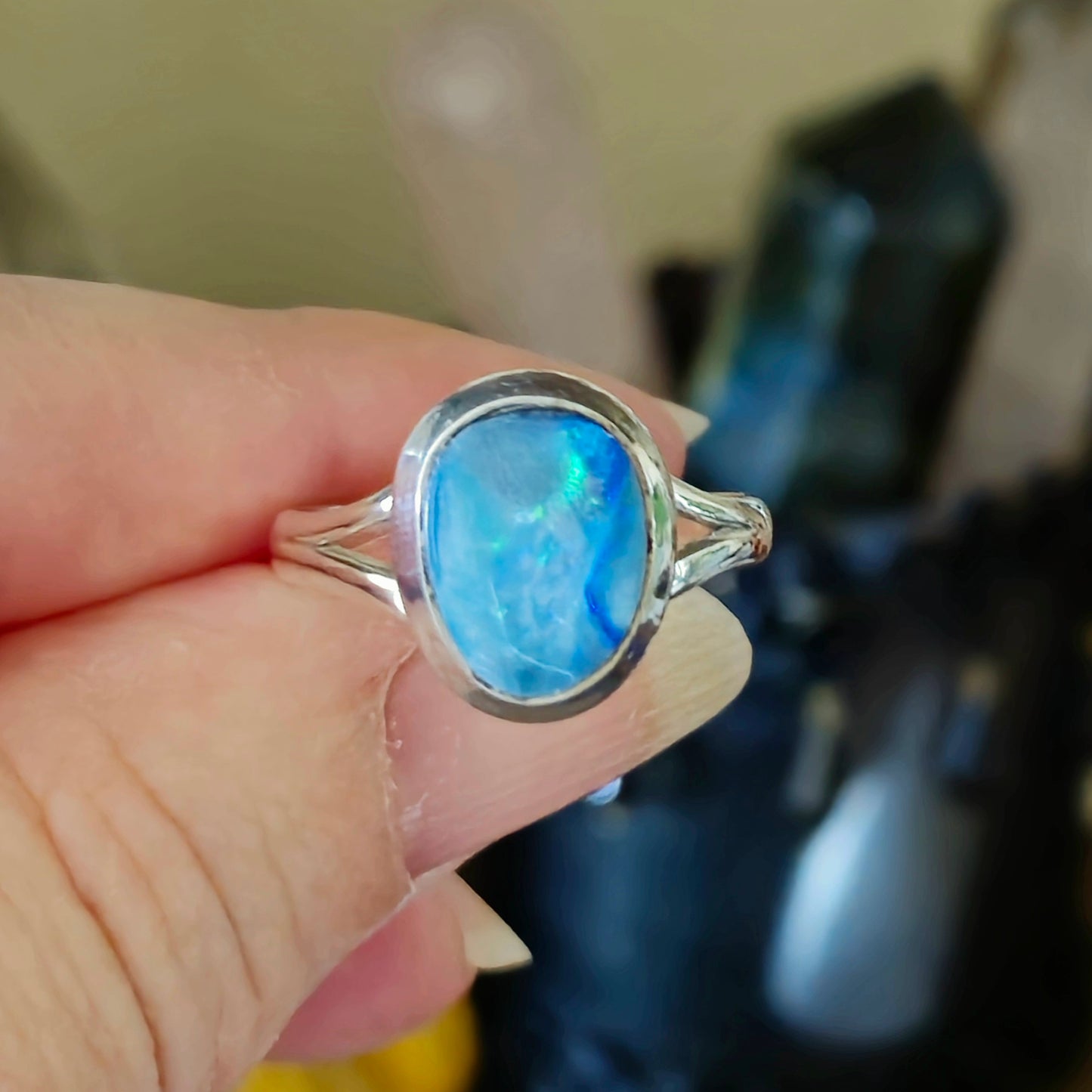 A person holding a blue opal ring with a metallic band, with a background of crystal stones.