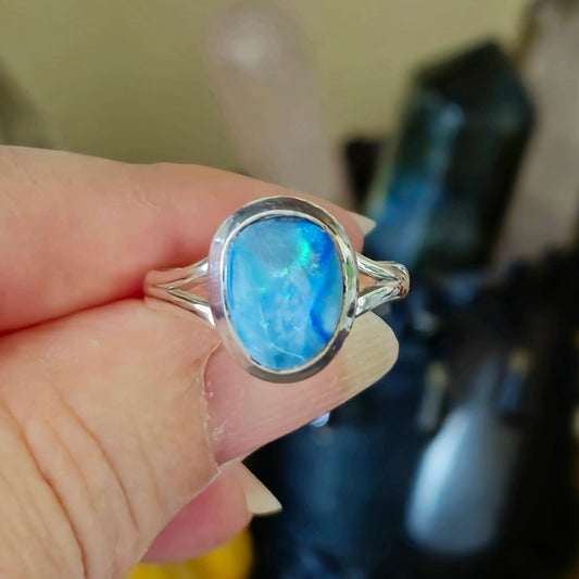 A person holding a blue opal ring with a metallic band, with a background of crystal stones.