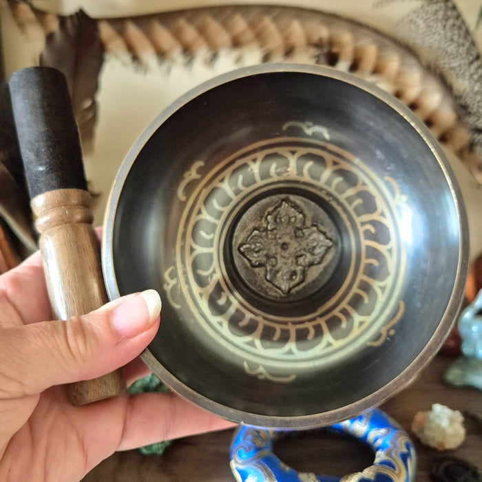 A person holding a brass singing bowl with a decorative design on the side, accompanied by a wooden mallet.