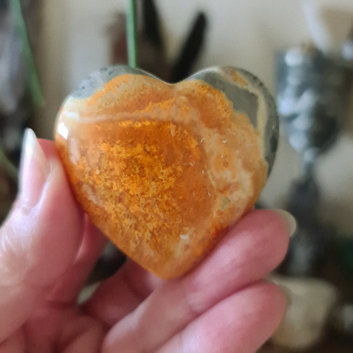 A person holding a polished heart-shaped Bumblebee Jasper stone with a reddish-orange hue.