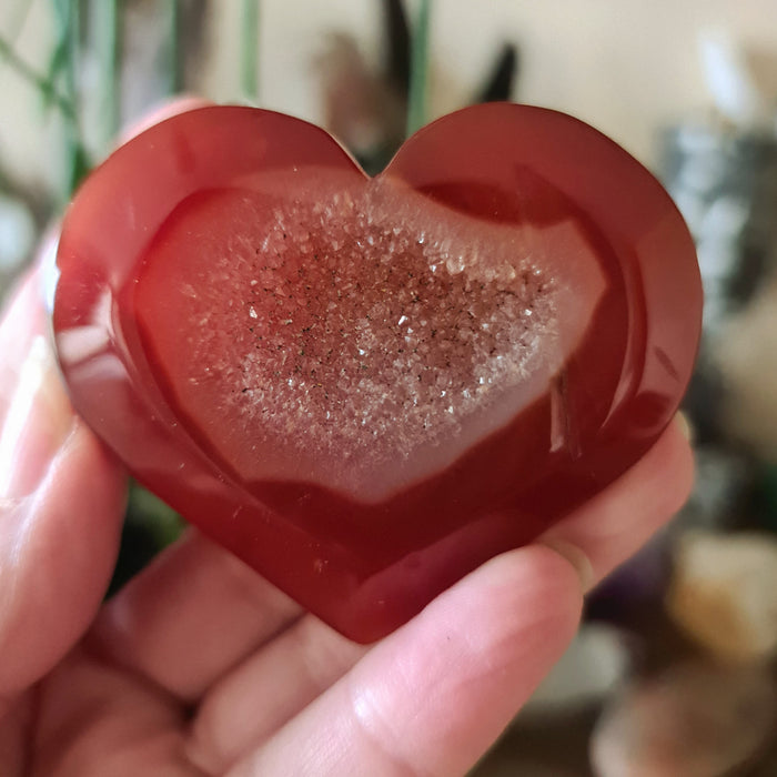 A red carnelian stone heart-shaped with a druzy cave effect, held in a person's hand.