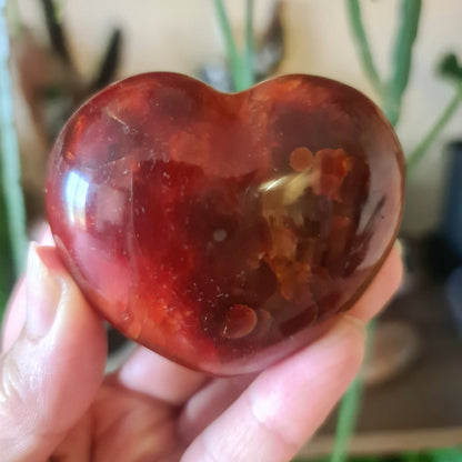 A hand holding a red carnelian heart-shaped stone with a smooth surface.
