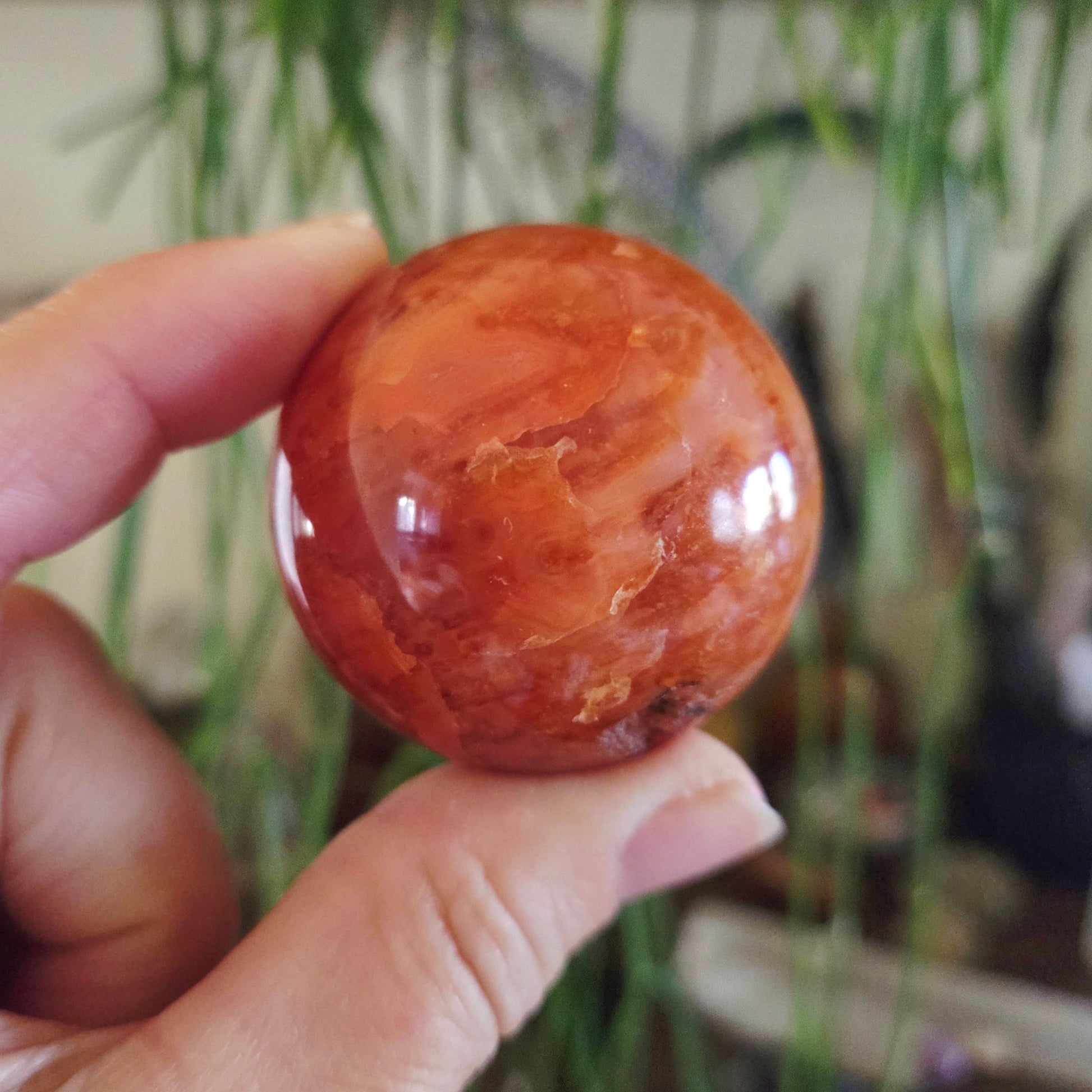 A hand holding a polished red and orange carnelian sphere with a smooth surface.
