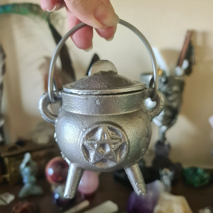 A small cast iron cauldron with a handle, displayed among other items, with a shiny metallic finish.