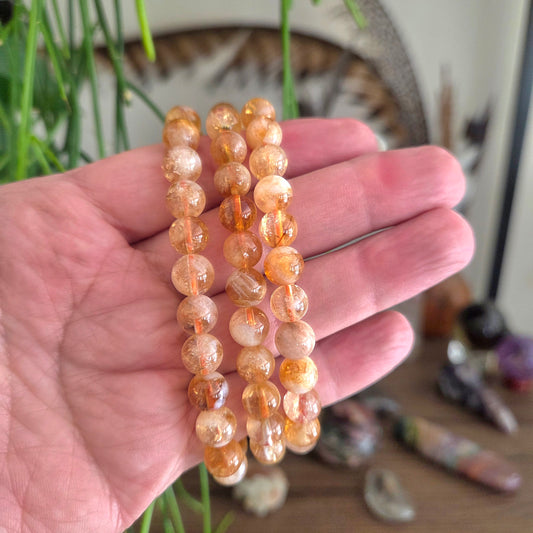 Three beaded stretch bracelets made of round, translucent orange and peach stones held in a person's hand, with a blurred background showing crystals and green plant stems.