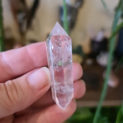 A hand holding a double-terminated clear quartz crystal with a polished surface, with a blurred background of greenery.