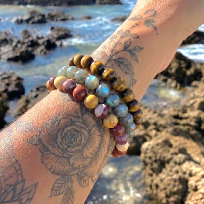 A close-up of a tattooed wrist wearing three beaded bracelets outdoors by the rocky ocean shore. The bracelets feature mixed stone colors including tiger eye brown, light blue, yellow-gold, maroon, and cream.