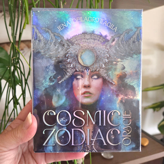 A hand holds a boxed card deck titled 'Cosmic Zodiac Oracle' by Jena Delagrotaglia. The box features an iridescent, cosmic design with a zodiac-themed illustration of a woman wearing a large ornate headdress against a galaxy background.