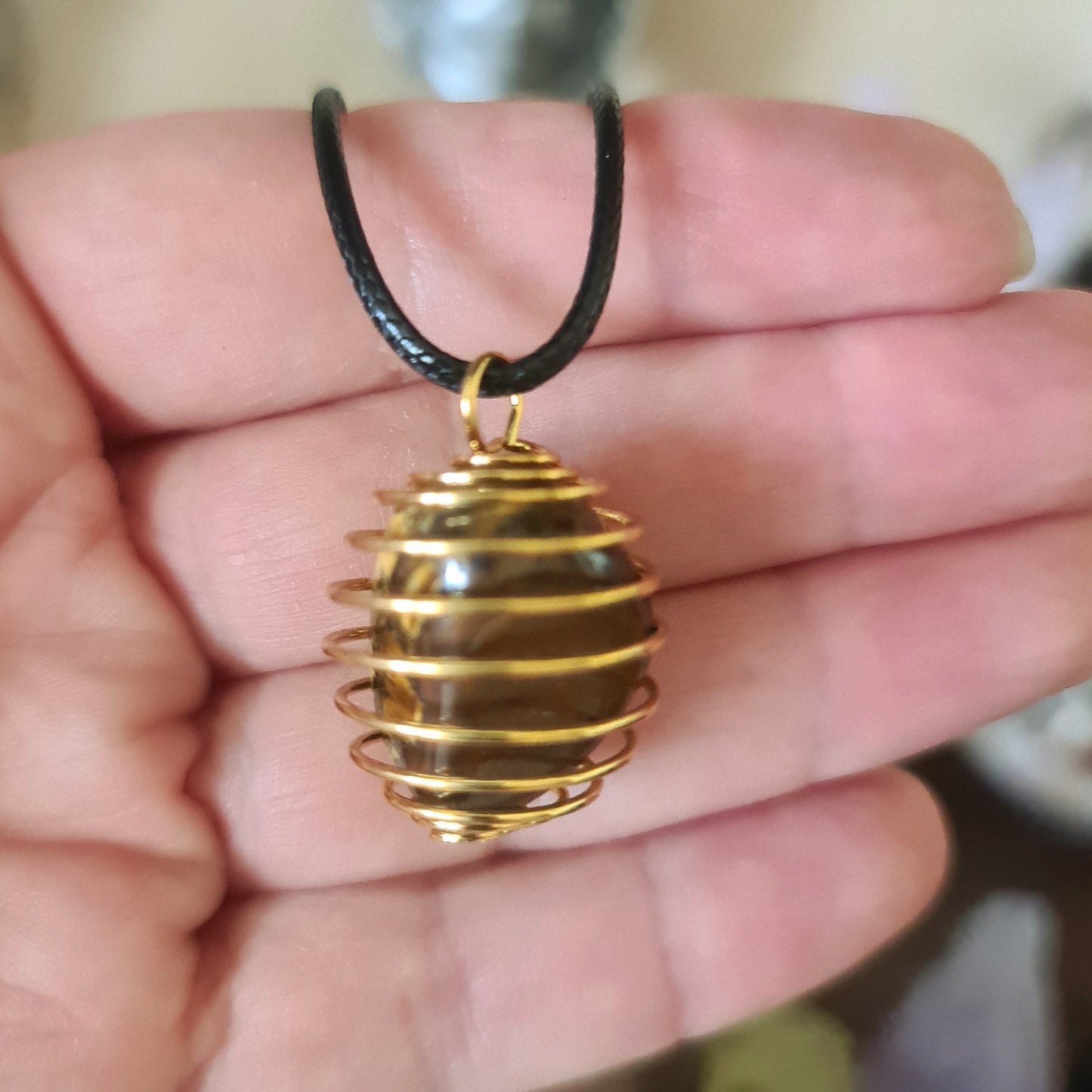 A gold-colored stainless steel tumbled stone holder in the shape of a cage, with a gemstone inside, held in a person's hand.