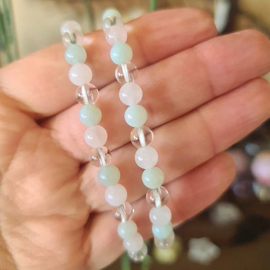 A hand holding a delicate beaded bracelet with a mix of clear, jade, and rose quartz crystals.