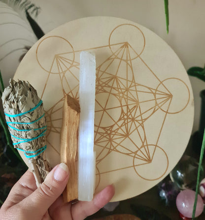 A hand holding a crystal grid board with a variety of crystals including a clear quartz point, rose quartz crystals, amethyst crystals, fluorite quartz crystals, a white sage torch, a Palo Santo stick, and a Selenite rod.