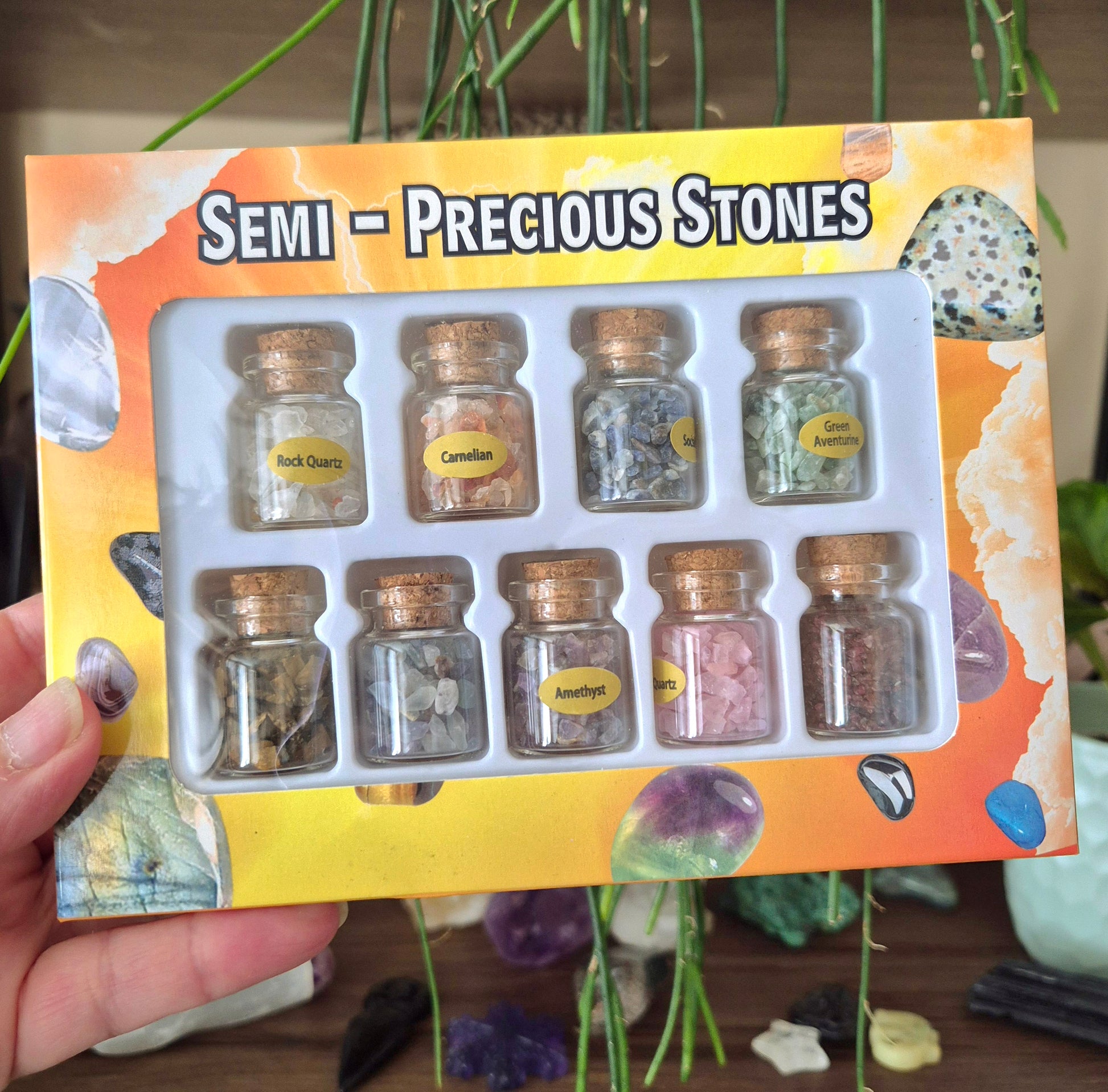 A pack of nine glass jars with crystal chips, labeled with different types of semi-precious stones, presented in a giftbox with a decorative background.