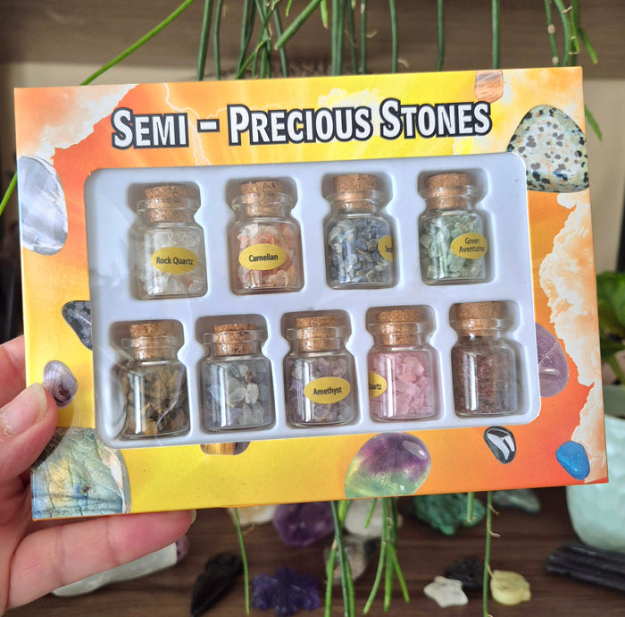 A pack of nine glass jars with crystal chips, labeled with different types of semi-precious stones, presented in a giftbox with a decorative background.