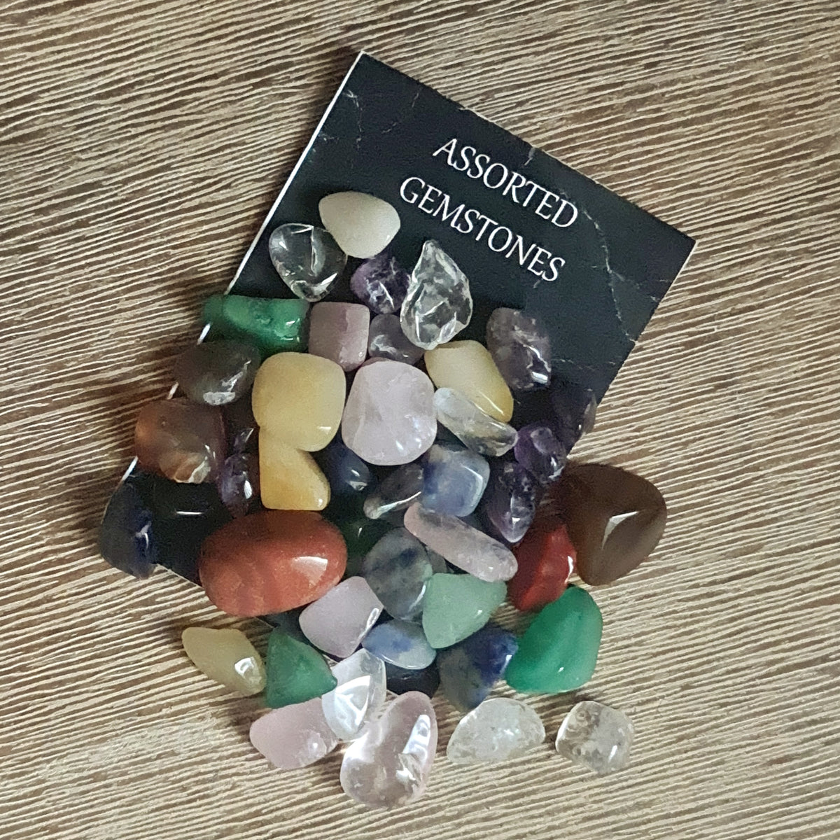 Assorted colorful gemstone chips and small tumbles displayed in front of a black card with the text 'Assorted Gemstones'.
