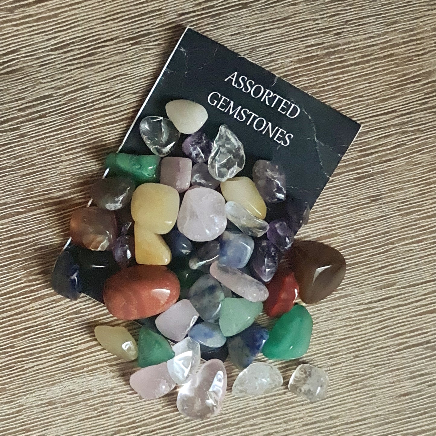 Assorted colorful gemstone chips and small tumbles displayed in front of a black card with the text 'Assorted Gemstones'.