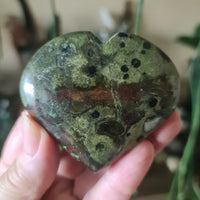 A heart-shaped polished stone with a green and brown color pattern, held in a person's hand.