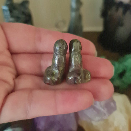This image displays a unique crystal figurine, shaped like a pair of penises, crafted from a stone known as Dragon Stone. The figurine is held in a person's hand, providing a sense of scale and demonstrating the textured, dark green surface of the stone.
