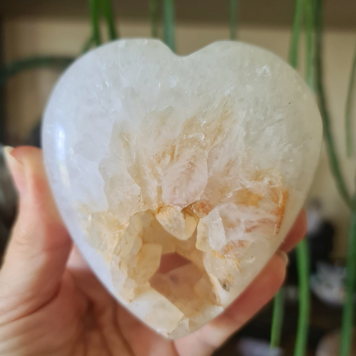 A polished druzy agate heart-shaped stone held in a person's hand, with a blurred green background suggesting indoor setting