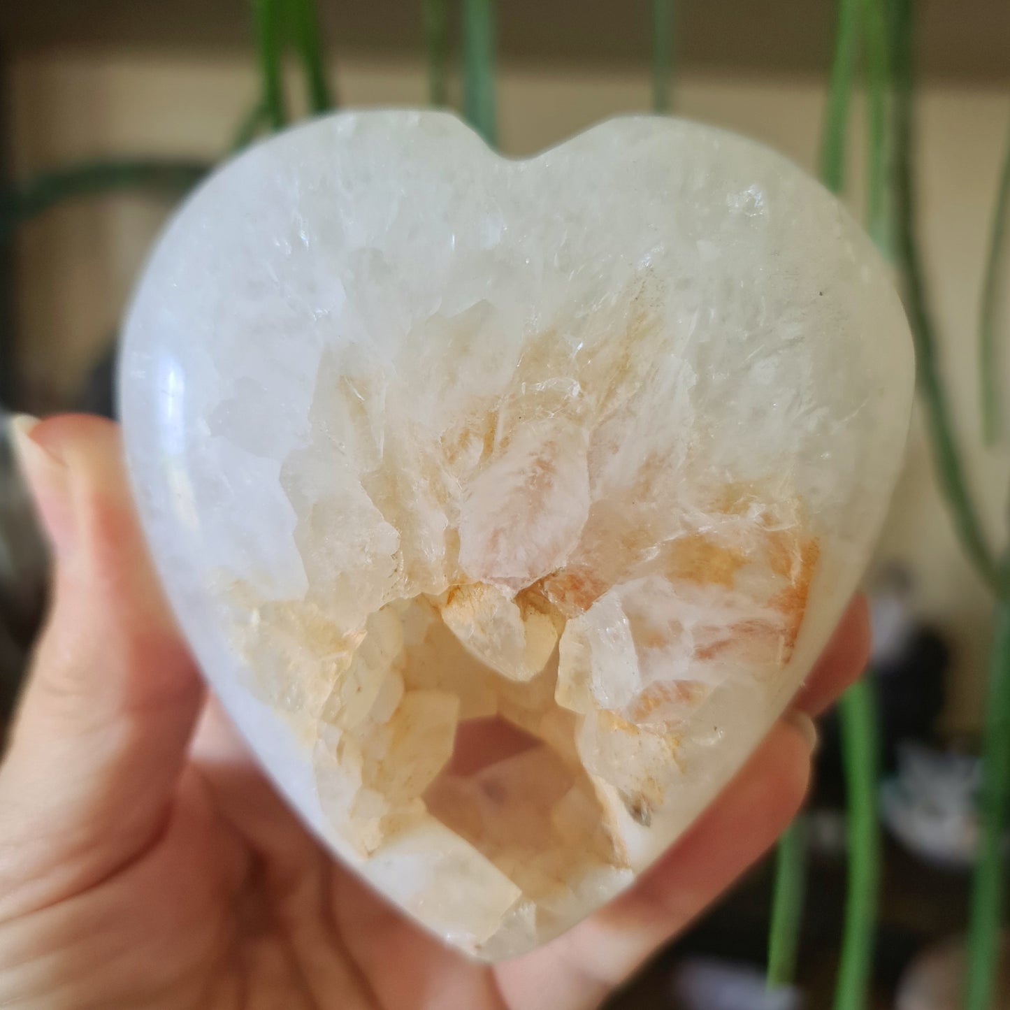 A polished druzy agate heart-shaped stone held in a person's hand, with a blurred green background suggesting indoor setting
