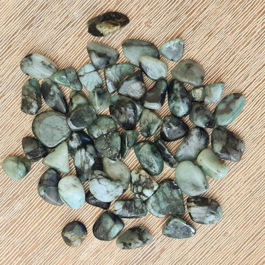 A collection of small emerald tumble stones with various shades of green and black, displayed on a beige surface.