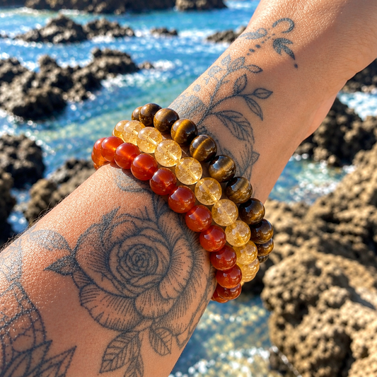 Three beaded stretch bracelets worn on a tattooed wrist, with a rocky beach and blue ocean in the background. The bracelets feature round beads in red-orange, translucent yellow, and brown-and-black tiger's eye.