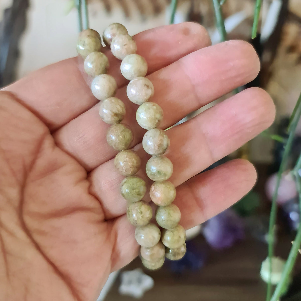 A hand holding a bracelet with polished, round epidote stones with varying shades of green.