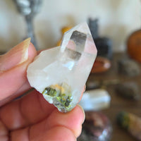 Hand holding a clear crystal with green inclusions against a blurred background