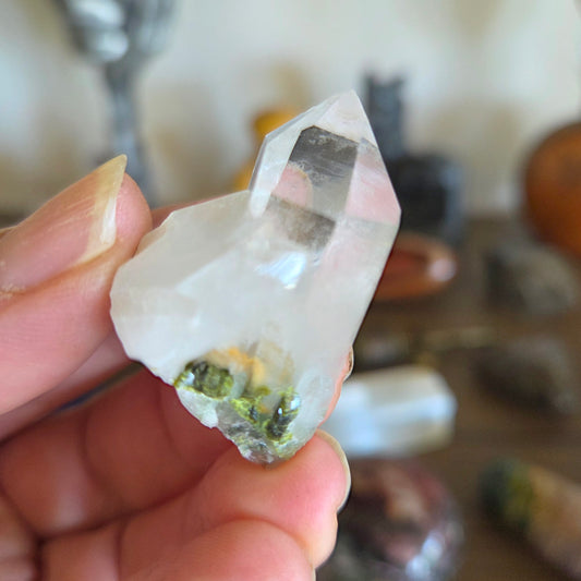 Hand holding a clear crystal with green inclusions against a blurred background