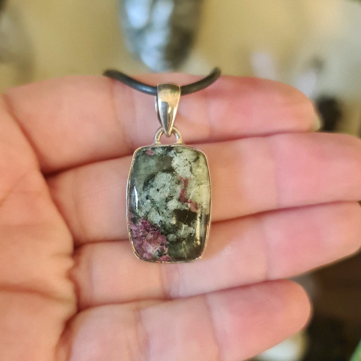 A hand holding a rectangular-shaped eudialyte pendant set in a silver mount, with a black cord.