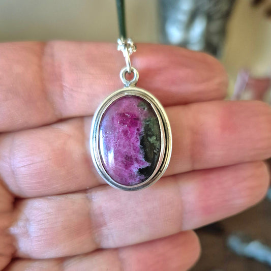 An oval-shaped eudialyte pendant held in hand, with a predominantly pink color and black inclusions, encased in a silver setting.