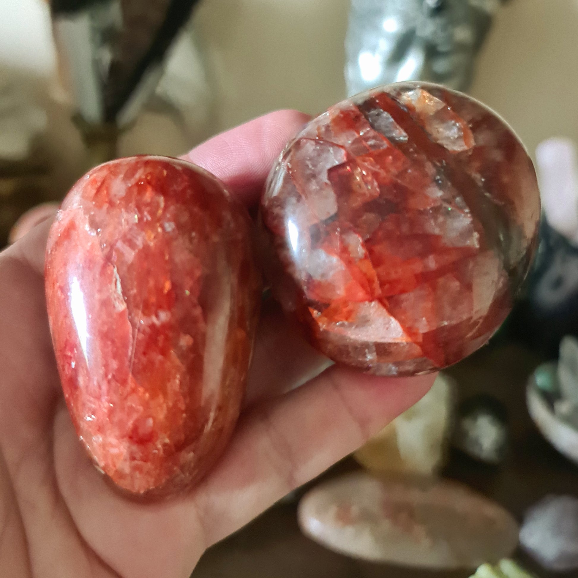 Two polished red Fire Quartz stones held in a person's hand, with a blurred background of various crystals and stones.