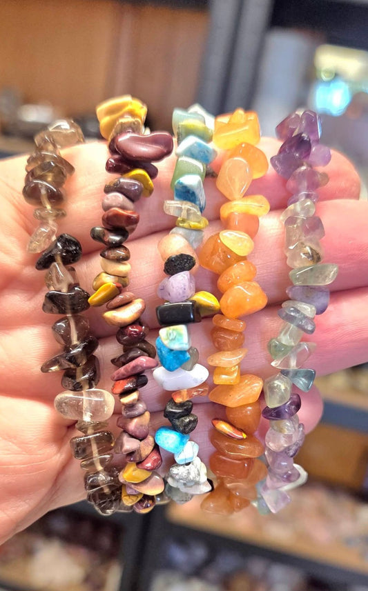 A hand holding six colorful tumbled stone chip bracelets in various colors including brown, yellow, blue, orange, purple, and green.