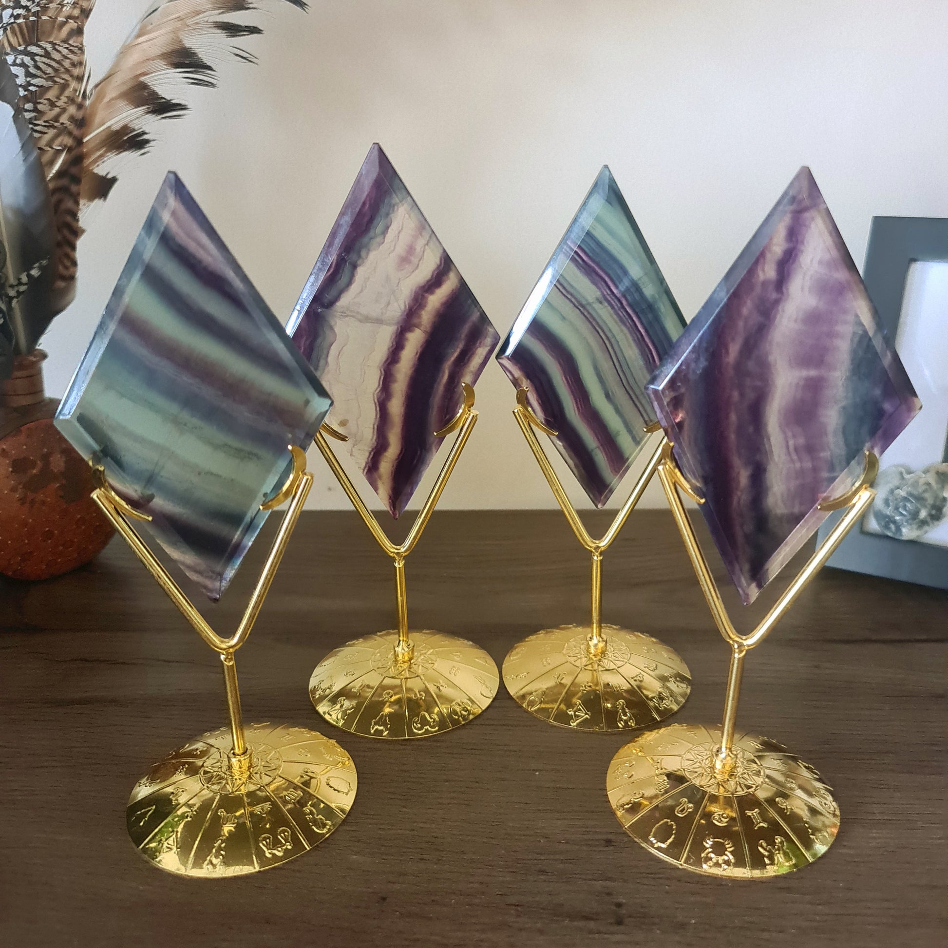 A fluorite crystal diamond carved into a diamond shape, mounted on a gold-colored zodiac sign stand, displayed on a wooden surface.