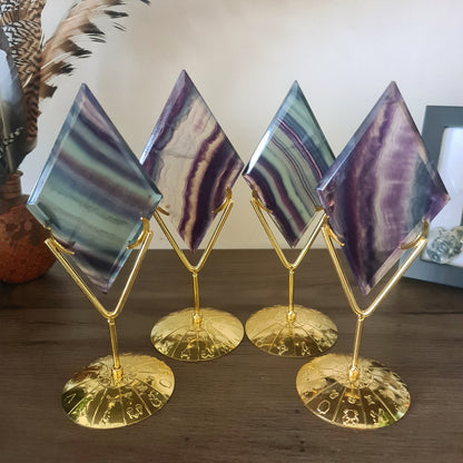 A fluorite crystal diamond carved into a diamond shape, mounted on a gold-colored zodiac sign stand, displayed on a wooden surface.