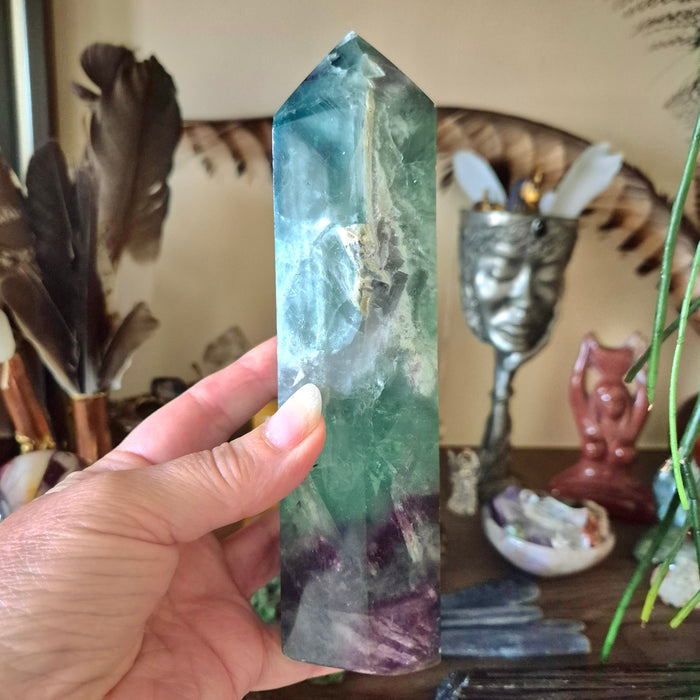 A hand holding a fluorite obelisk tower with a green and purple color scheme, placed against a backdrop of various decorative items.