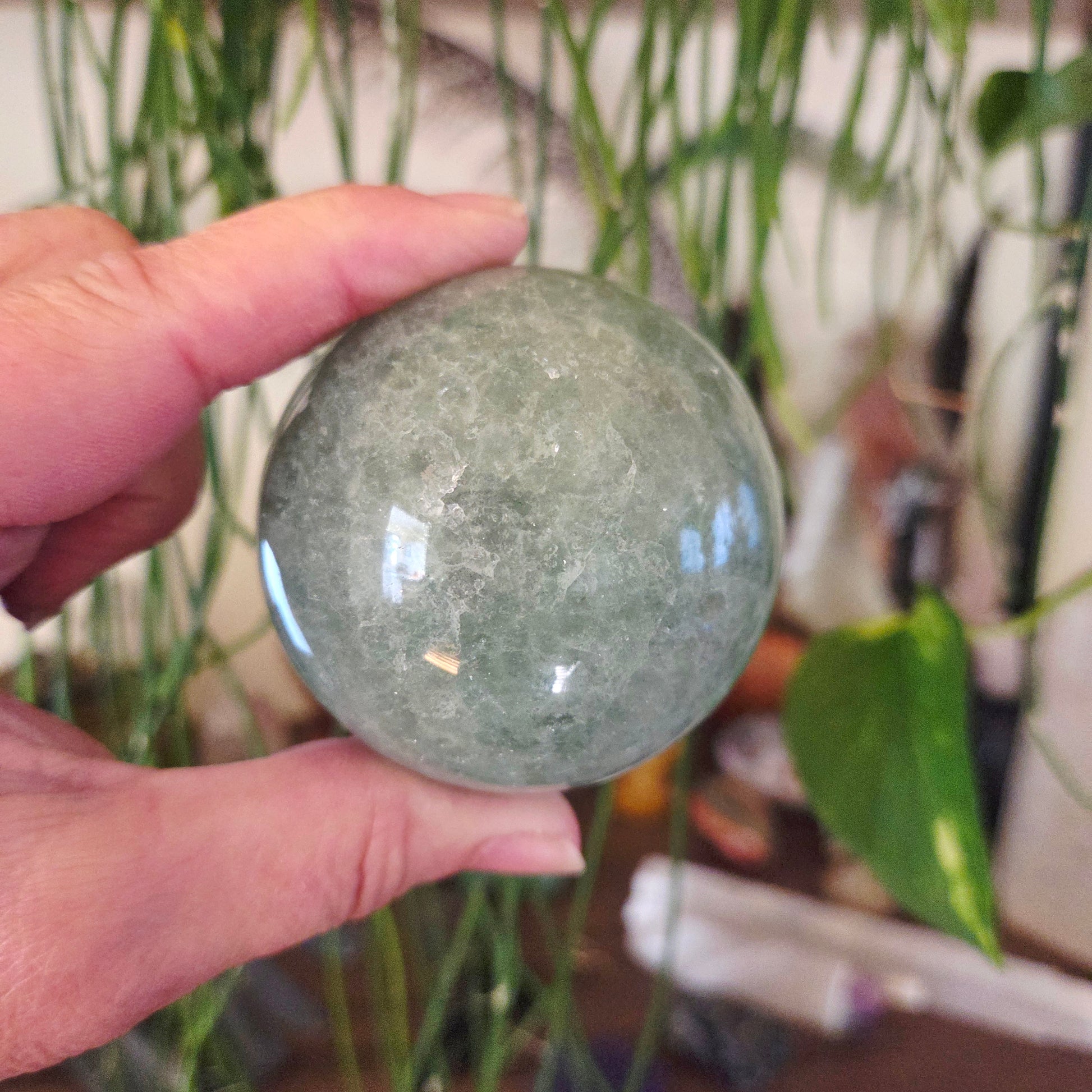A hand holding a green fluorite sphere with a plant in the background.