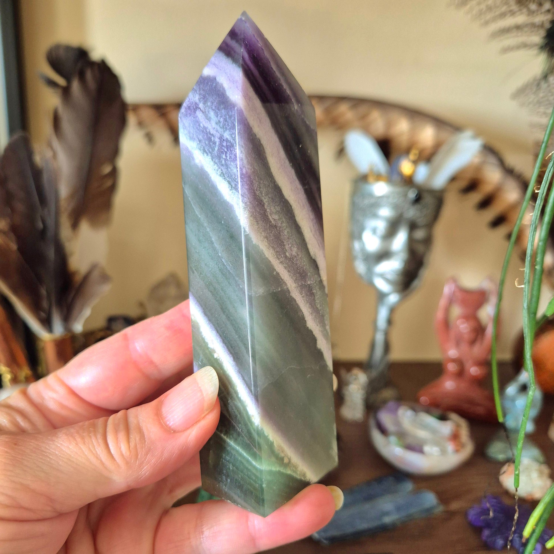 A hand holding a fluorite crystal with a purple and green color pattern, displayed against a backdrop of various decorative items.